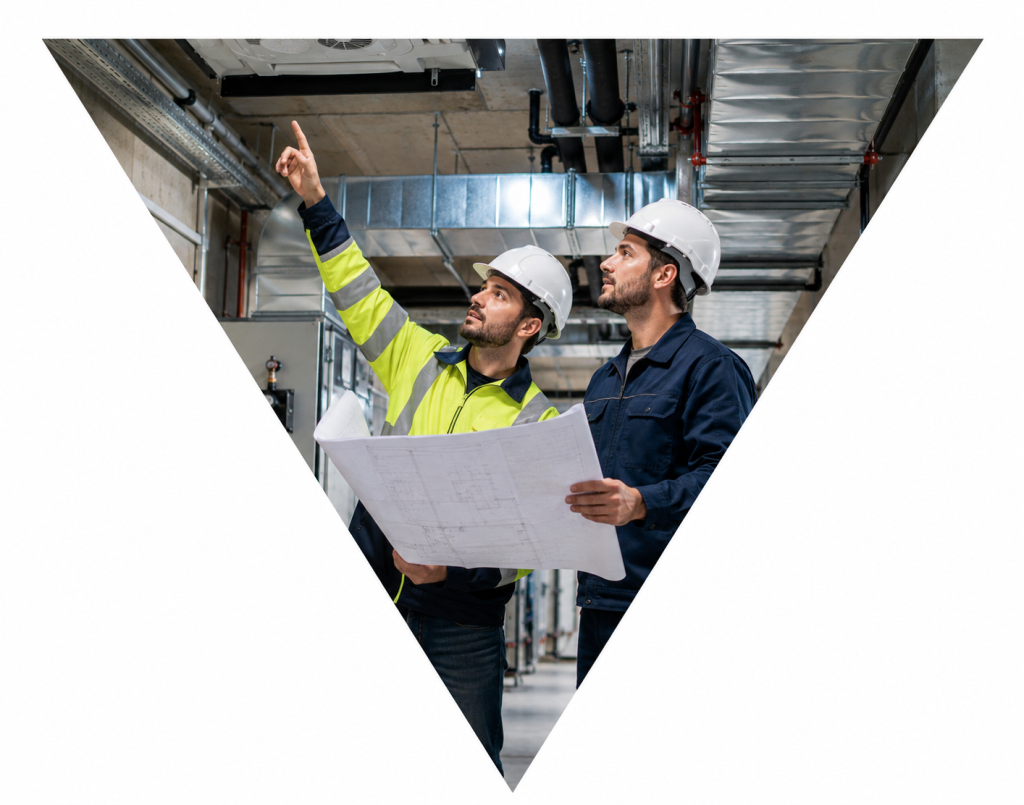 Dos técnicos con casco revisando planos en una instalación interior con conductos y sistemas técnicos.